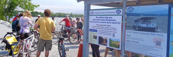 Photo: Aude Lochet Group of people with bikes on Lake Champlain causeway next to Little Lake Lesson sign